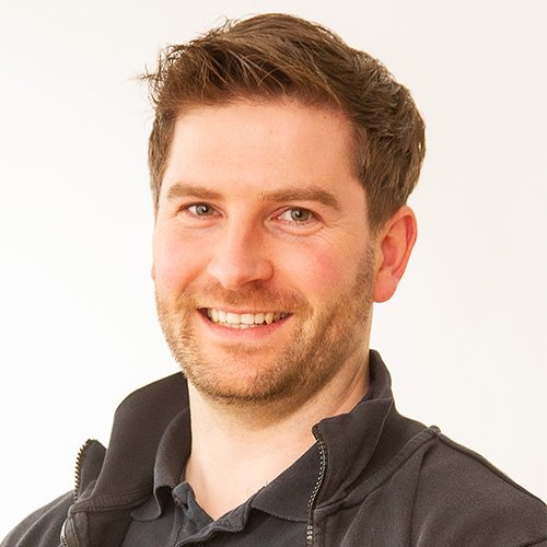 Der Mann hat einen freundlichen Gesichtsausdruck und trägt einen schwarzen Pullover, während er in die Kamera schaut.