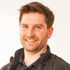 Der Mann hat einen freundlichen Gesichtsausdruck und trägt einen schwarzen Pullover, während er in die Kamera schaut.