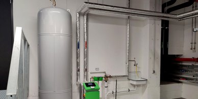 Ein großer, grauer Wassertank steht an einer Wand, umgeben von silbernen Rohren und einem grünen Steuergerät.