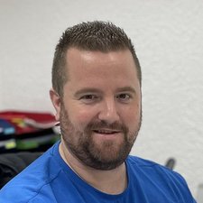 Ein lächelnder Mann mit kurzem, gestutztem Bart trägt ein blaues T-Shirt und sitzt in einem hellen Raum mit verschwommenem Hintergrund.