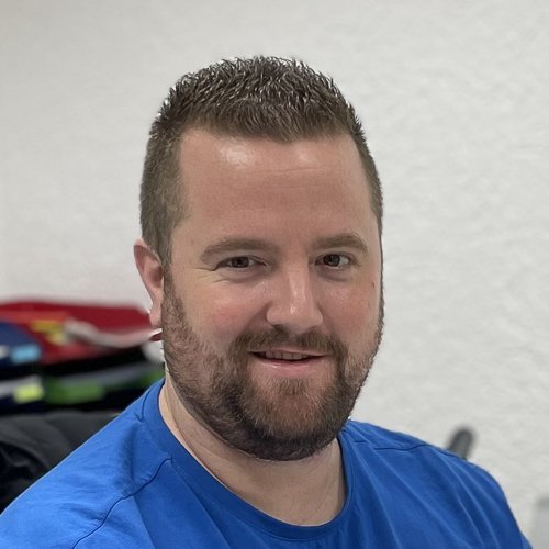 Ein lächelnder Mann mit kurzem, gestutztem Bart trägt ein blaues T-Shirt und sitzt in einem hellen Raum mit verschwommenem Hintergrund.