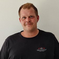 Der Mann trägt ein schwarzes T-Shirt mit einem Logo und hat einen Ohrstecker im linken Ohr.