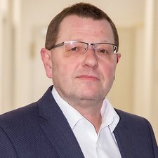 Ein Mann mit Brille und Anzug schaut direkt in die Kamera, mit einem neutralen Gesichtsausdruck in einem hellen Raum.