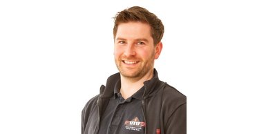 Ein lächelnder Mann mit braunem Haar trägt ein schwarzes Poloshirt und steht vor einem weißen Hintergrund.