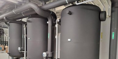 Drei große, schwarze Behälter stehen nebeneinander in einem modernen Raum, umgeben von Rohren und technischen Geräten.