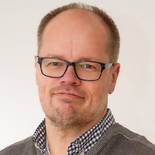 Der Mann hat eine entspannte Ausstrahlung, trägt eine karierten Hemdkragen unter einem grauen Pullover und schaut direkt in die Kamera.