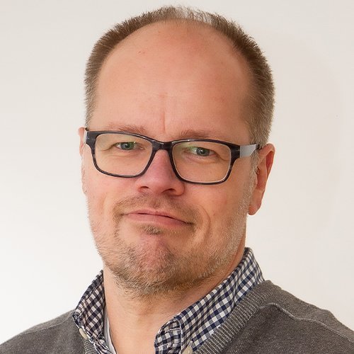Der Mann hat eine entspannte Ausstrahlung, trägt eine karierten Hemdkragen unter einem grauen Pullover und schaut direkt in die Kamera.